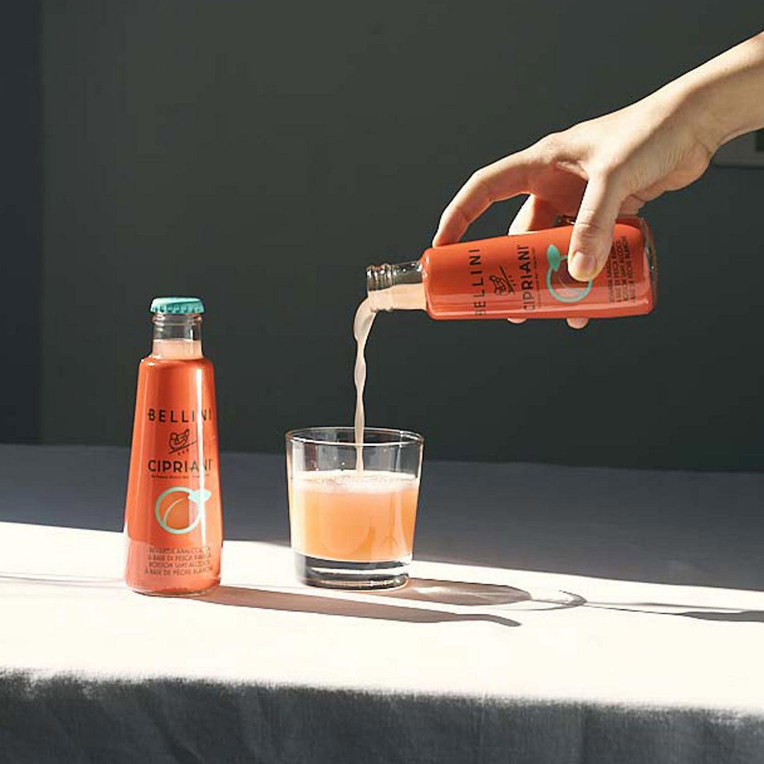 Hand pouring Cipriani aperitif into glass beside bottle on neutral background