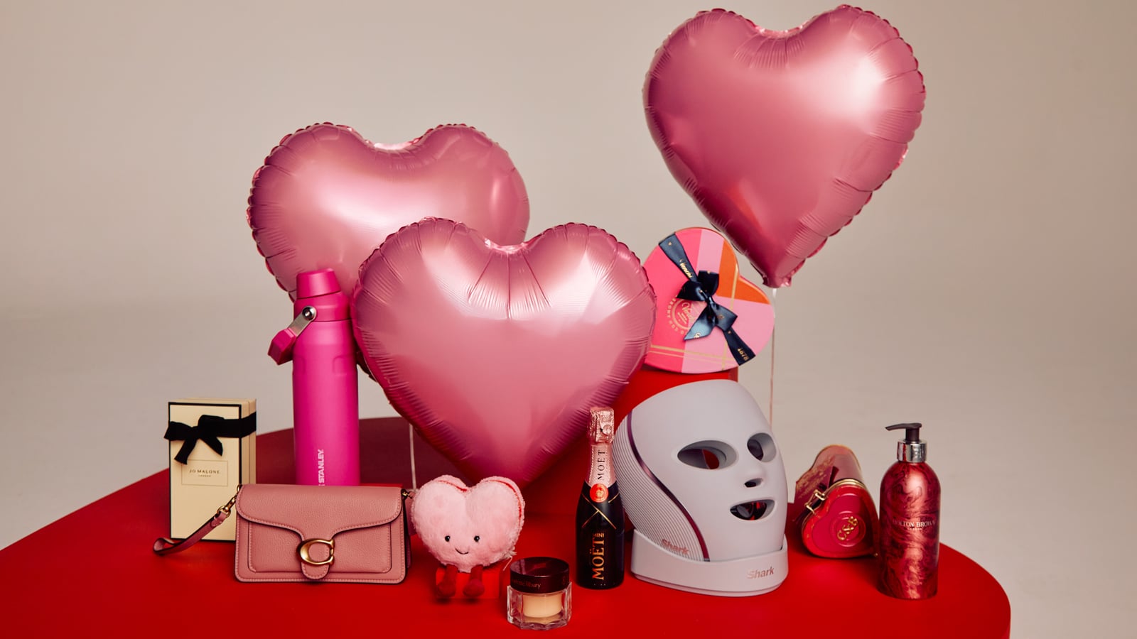 A collection of items on a red surface, including pink heart-shaped balloons, a purse, gift boxes, a face mask device, and various cosmetics.