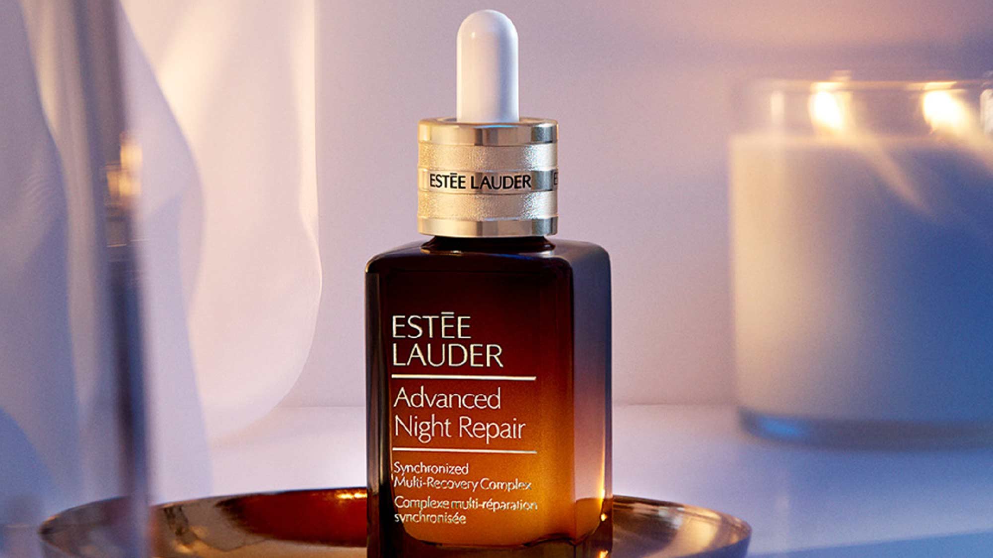 A bottle of Estée Lauder Advanced Night Repair serum on a golden tray, with a lit candle in the background.