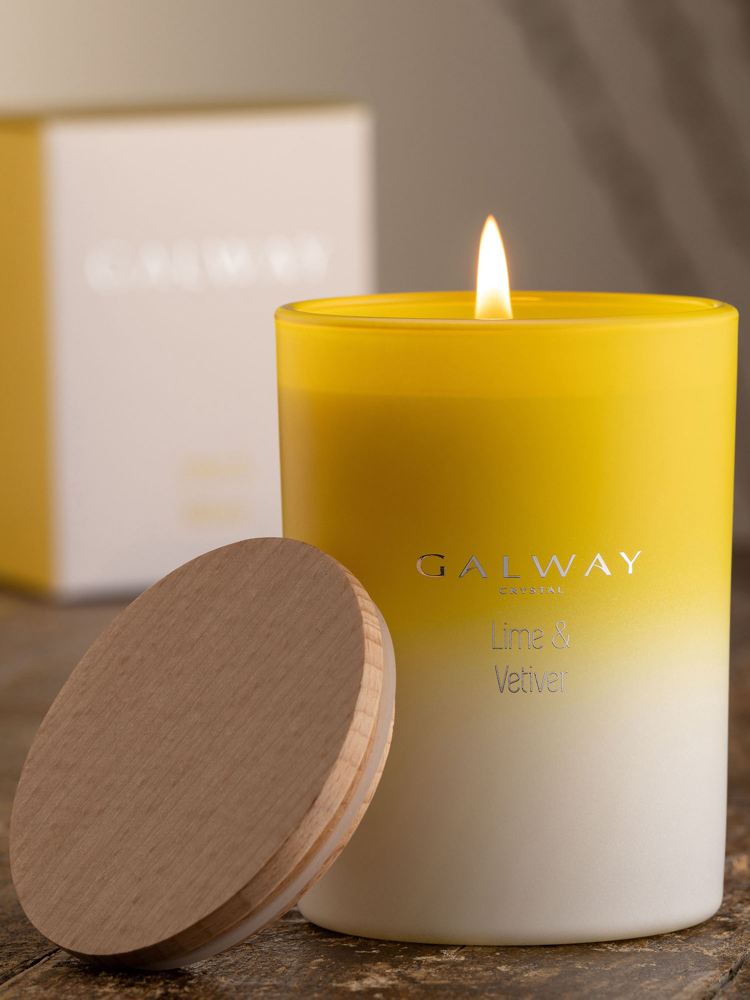 A yellow and white candle labeled 'Lime & Vetiver' burns on a wooden surface, next to its wooden lid, with its box in the background.