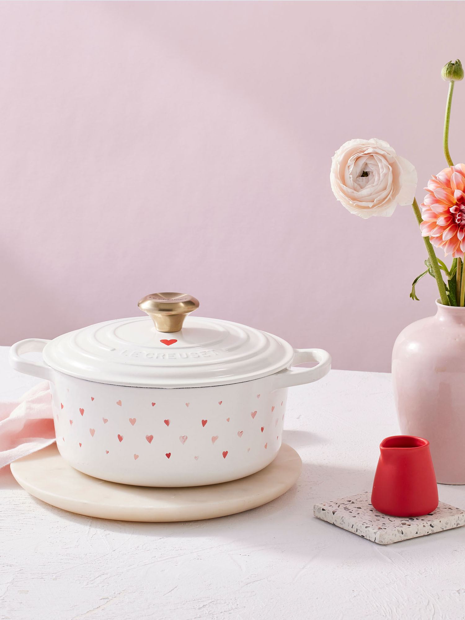 White pot with pink heart pattern, pink vase with flowers, small red vase, and coaster on a white table against a pink background.