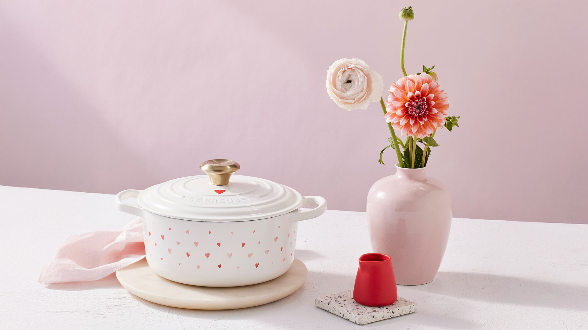 White pot with pink heart pattern, pink vase with flowers, small red vase, and coaster on a white table against a pink background.