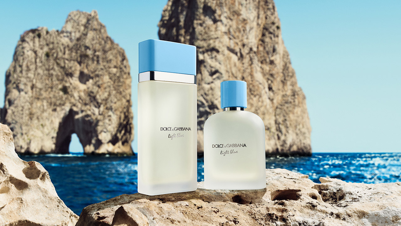Two Dolce & Gabbana Light Blue perfume bottles are set against a scenic ocean backdrop with rocky formations.