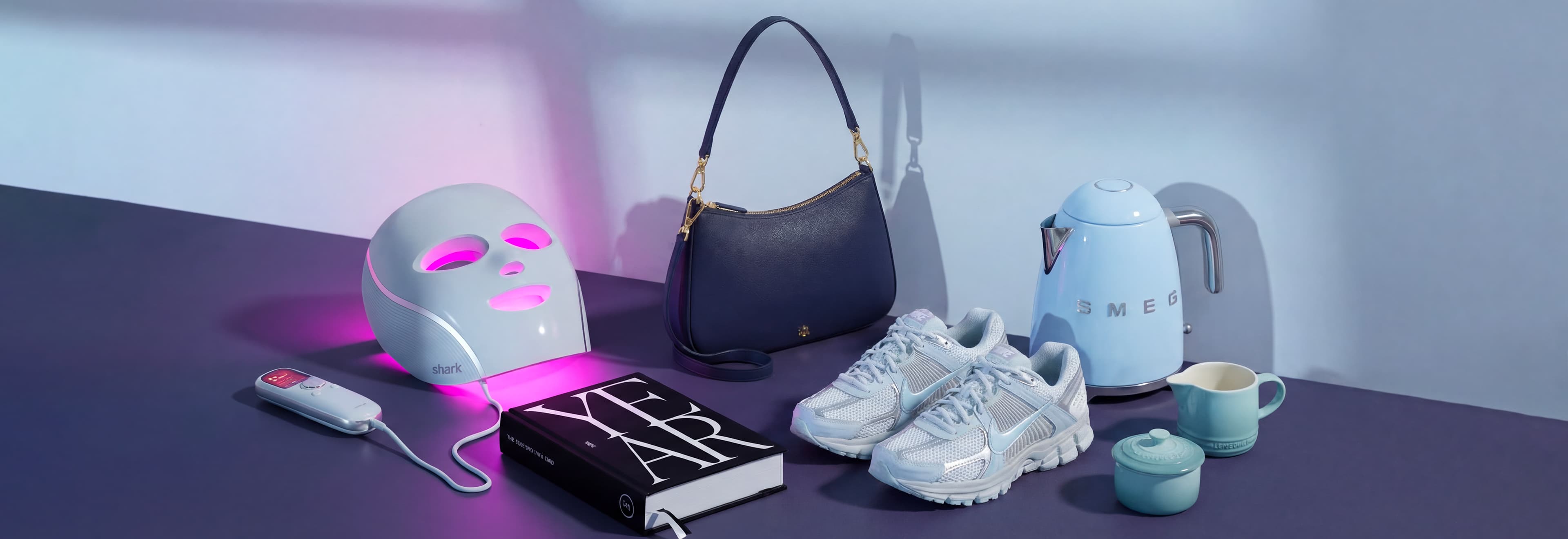 A collection of items including an LED face mask, book, handbag, sneakers, kettle, and teacups on a table.