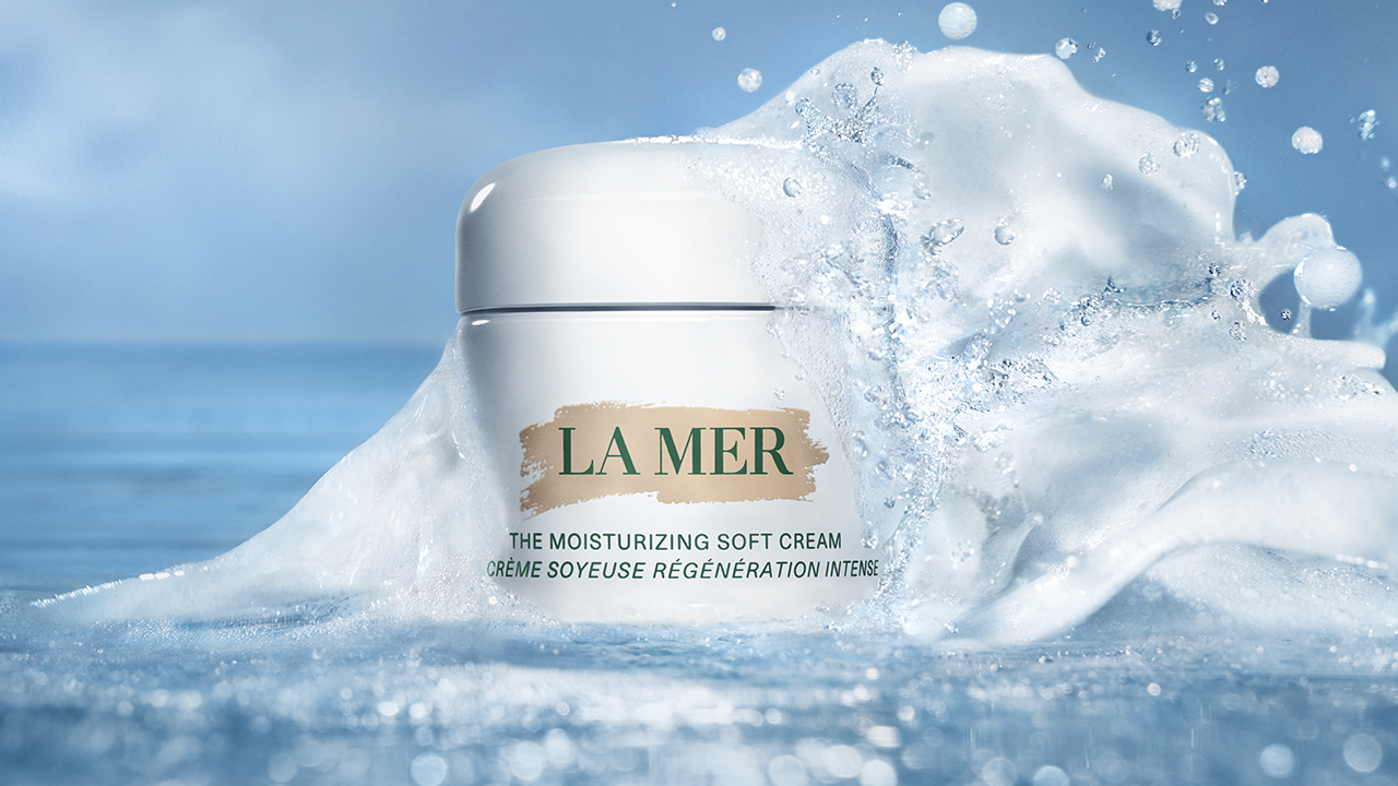 A jar of La Mer moisturizing soft cream surrounded by splashes of water against a blue background.