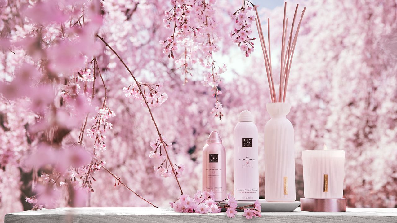 Pink beauty products on a table surrounded by cherry blossoms, featuring a candle and a reed diffuser.