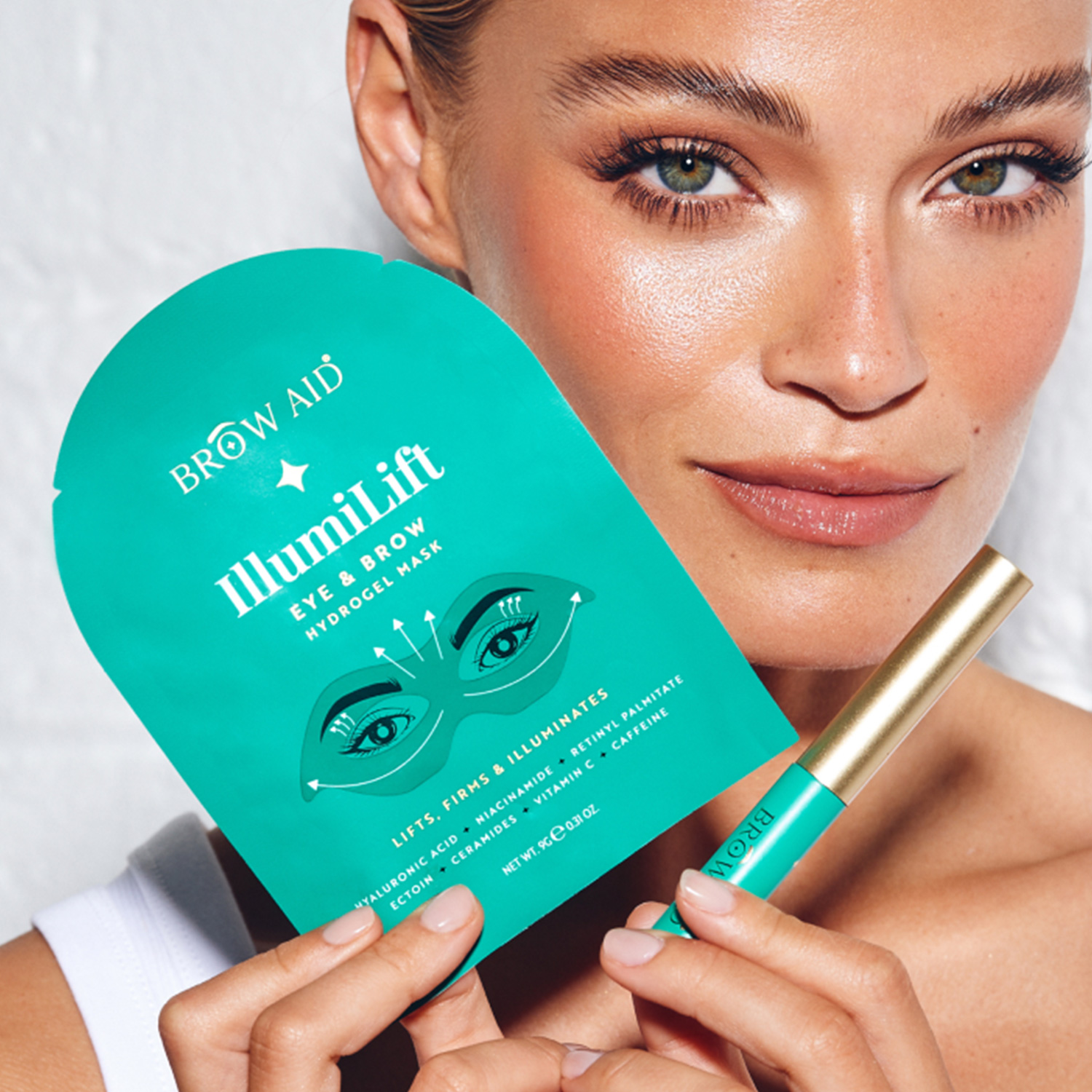 A person holds a teal package labeled 'IllumiLift Eye & Brow Hydrogel Mask' and a matching pen. They have flawless makeup and light blue eyeshadow.