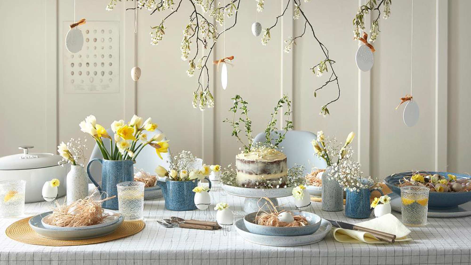 Elegant spring table setting with daffodils, a cake, and hanging decorations. Plates feature decorative nests.