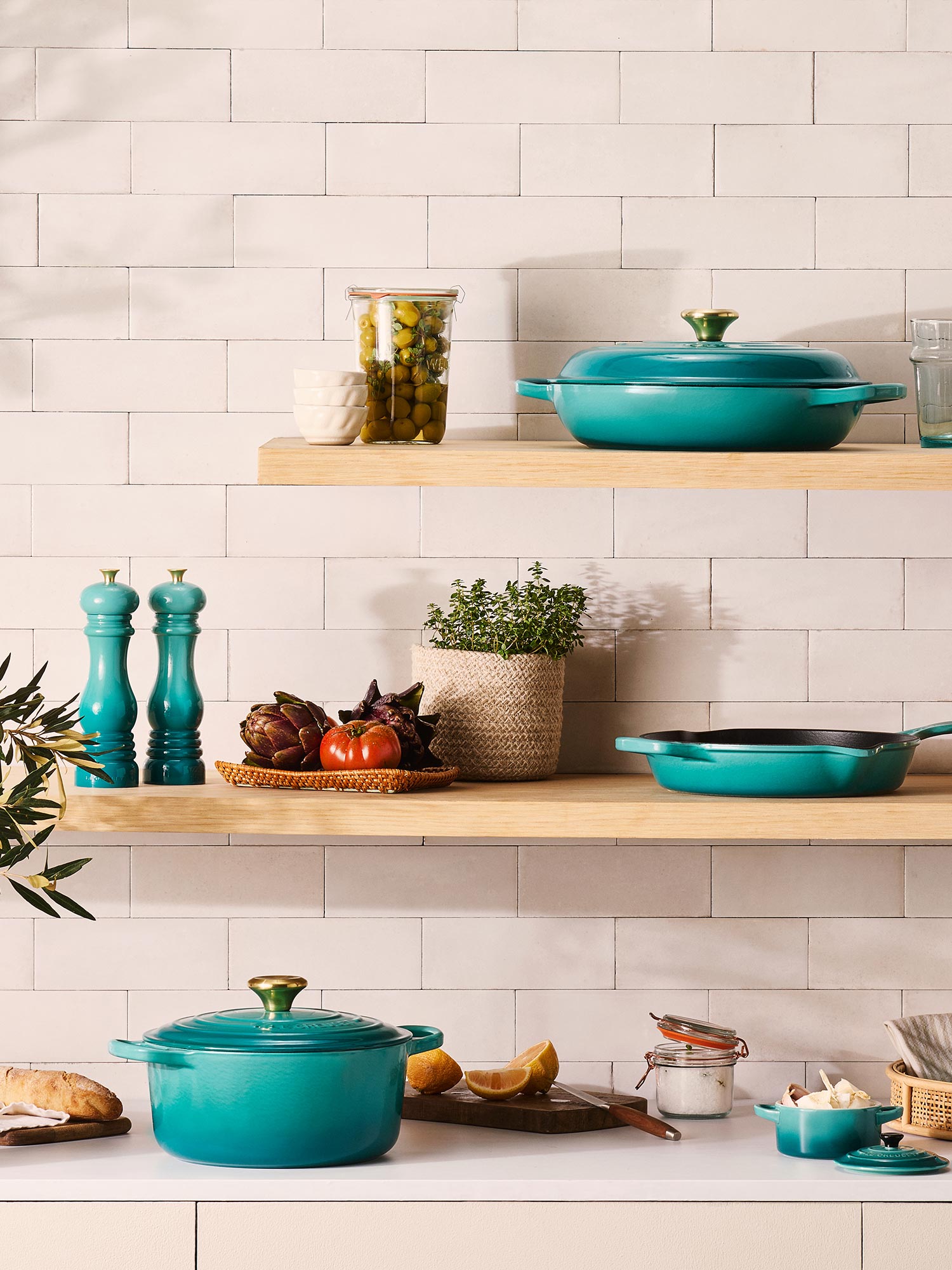 Kitchen shelf with turquoise cookware, potted herbs, vegetables, and condiments against a tiled wall.