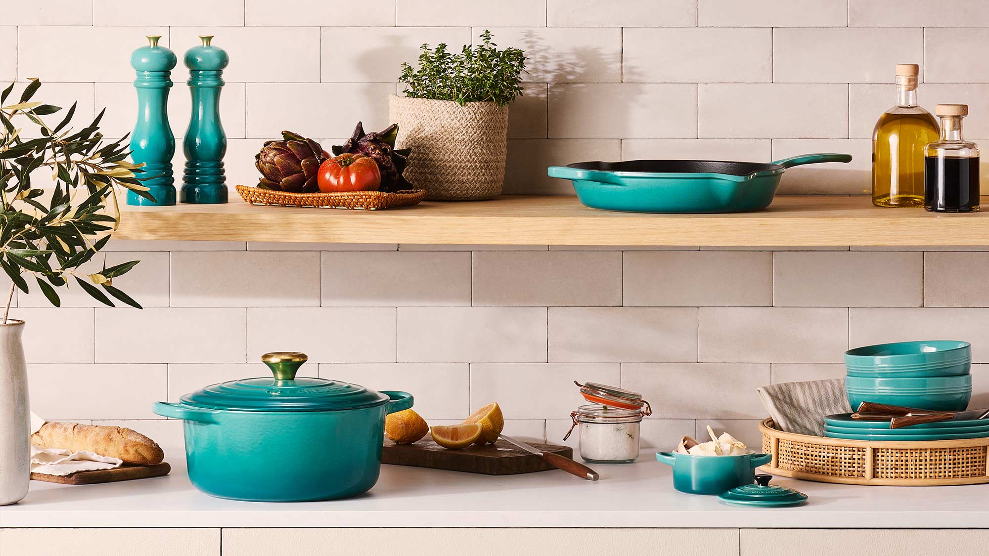 Kitchen shelf with turquoise cookware, potted herbs, vegetables, and condiments against a tiled wall.