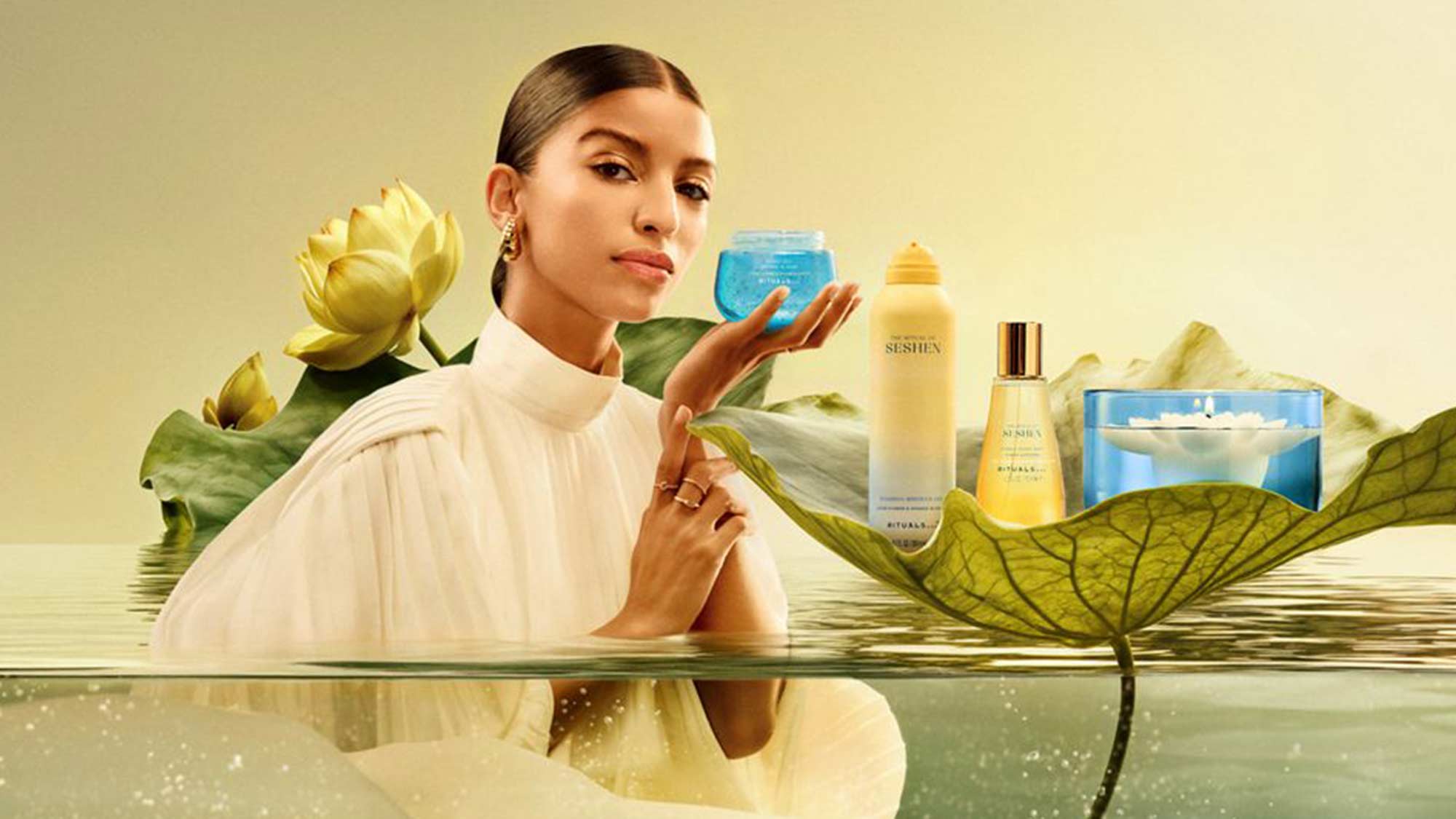 A woman in a white dress stands in water, holding a blue jar. Skincare products are displayed on a large leaf.
