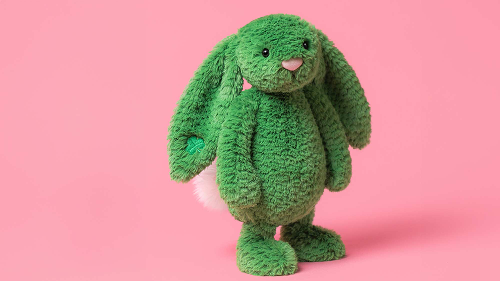A green plush bunny with long ears and a white tail, standing against a pink background.