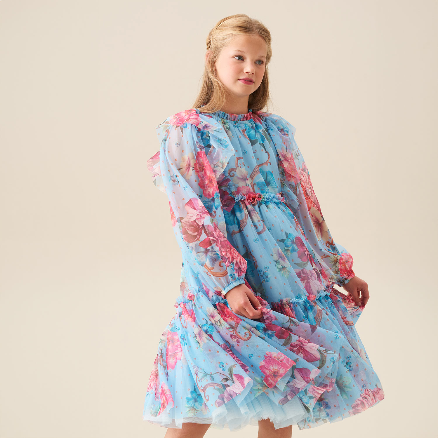 A young girl wearing a blue floral dress with pink flowers stands against a plain backdrop.