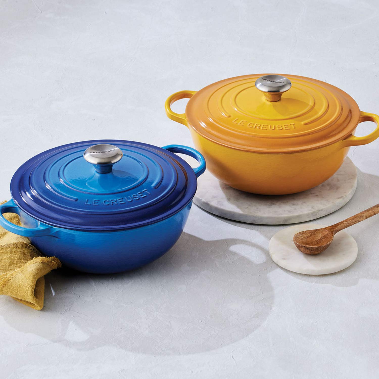 Two Le Creuset Dutch ovens, one blue and one yellow, with a wooden spoon and a cloth on a light surface.