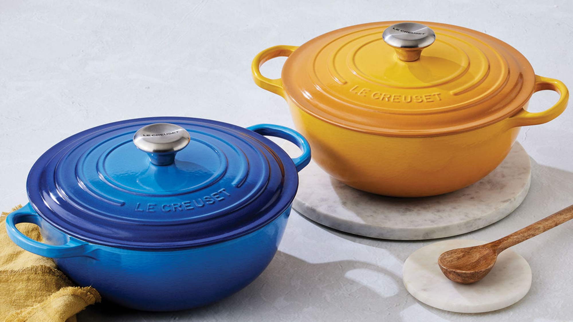Two Le Creuset Dutch ovens, one blue and one yellow, with a wooden spoon and a cloth on a light surface.
