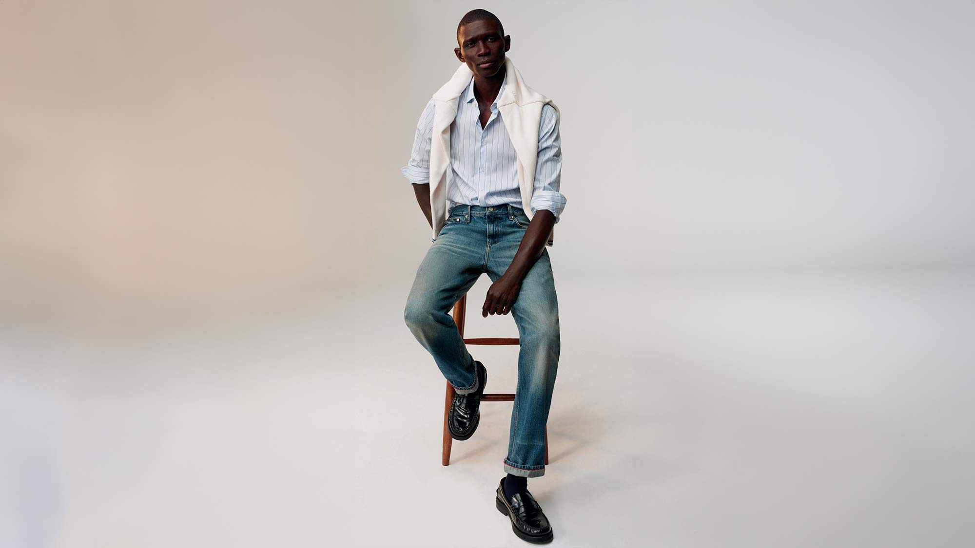 A person sits on a stool wearing a striped shirt, jeans, and loafers, with a sweater draped over their shoulders.