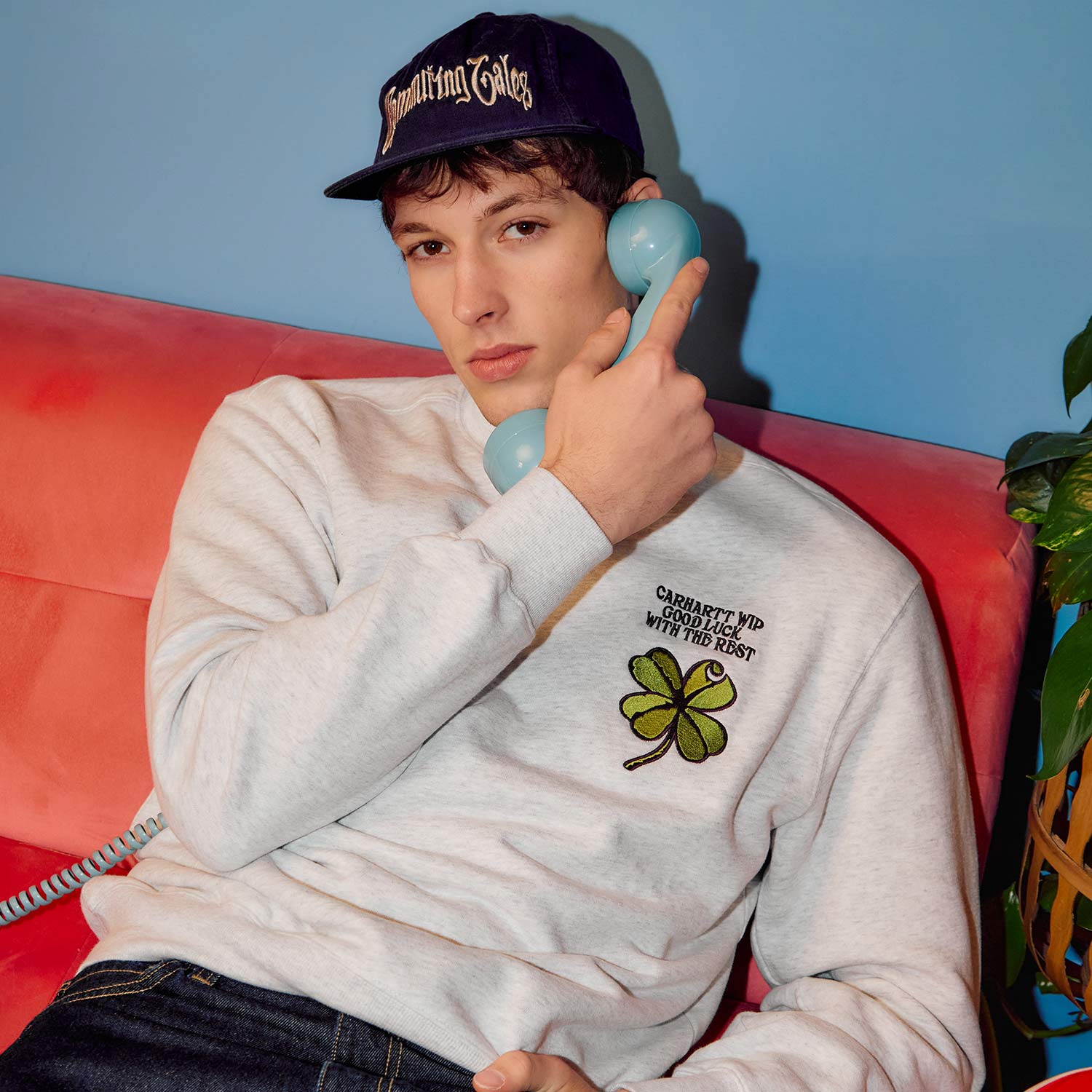 Person sitting on a pink couch, holding a blue retro phone to their ear, wearing a cap and sweatshirt with a clover design.