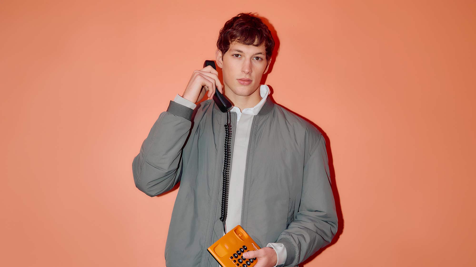 A person in a gray jacket holds an orange retro phone against an orange background.