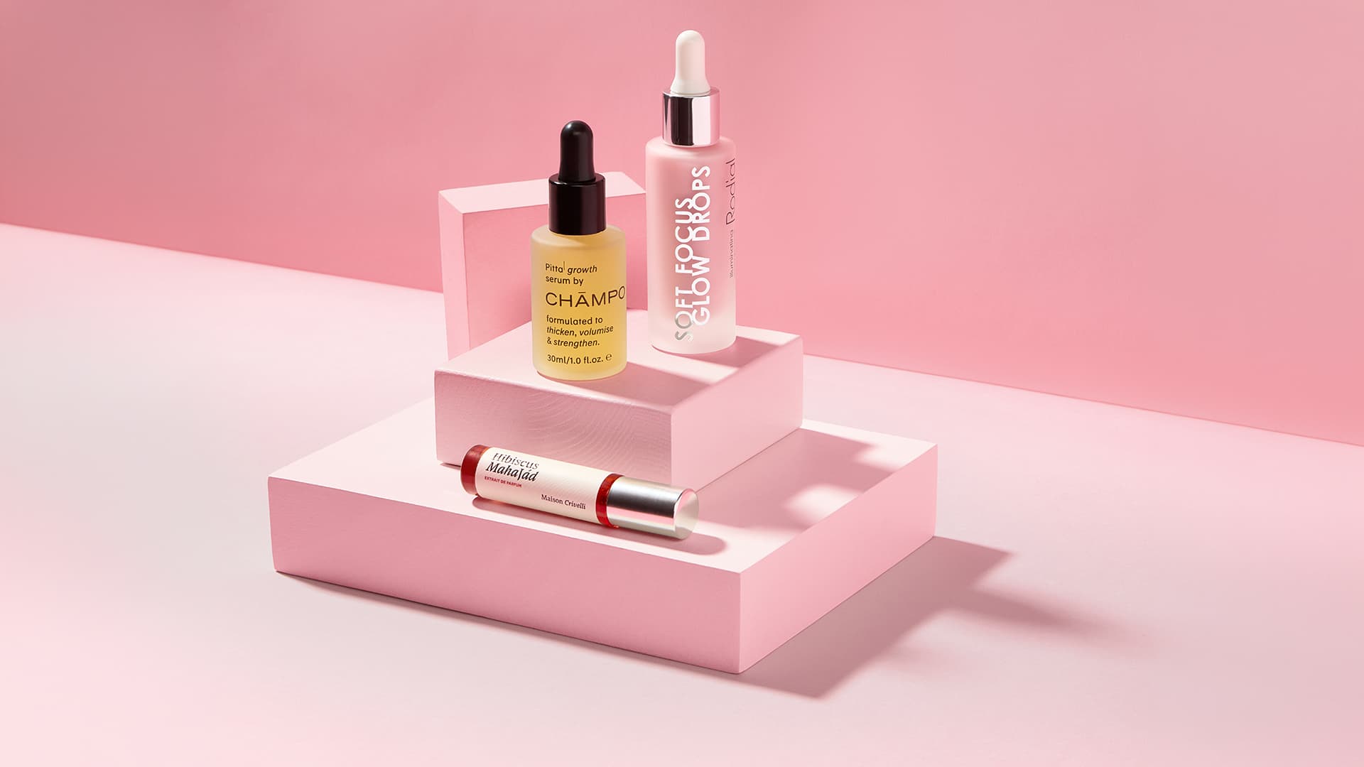 Three skincare products on pink platforms: a dropper bottle of yellow serum, a clear dropper bottle, and a tube of cream.