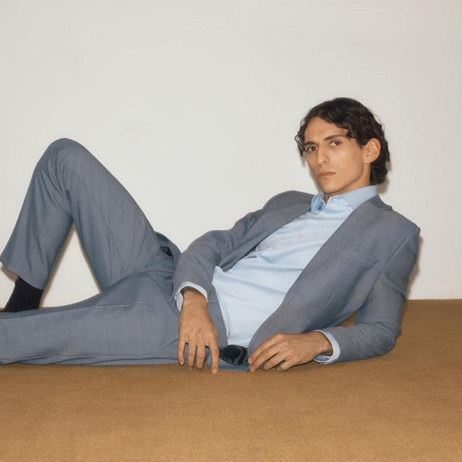 A person in a gray suit lies on the floor against a white background, looking at the camera.