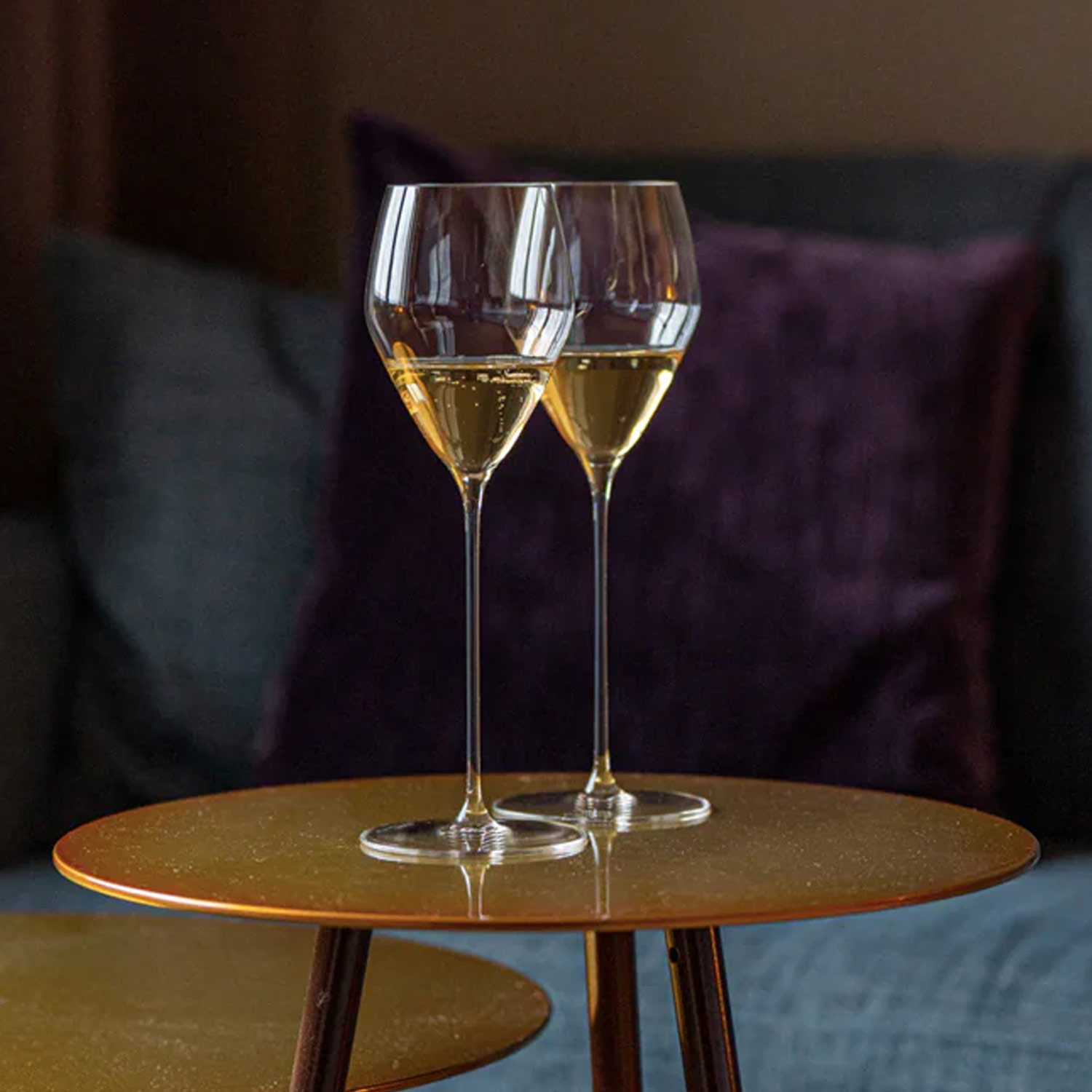 Two glasses of champagne on a small round table, with a sofa and purple pillow in the background.