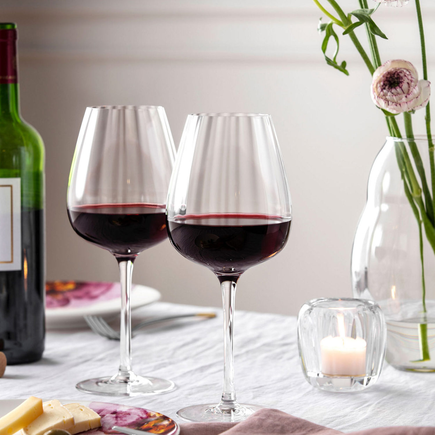 Two glasses of red wine on a table with cheese, a candle, and flowers.