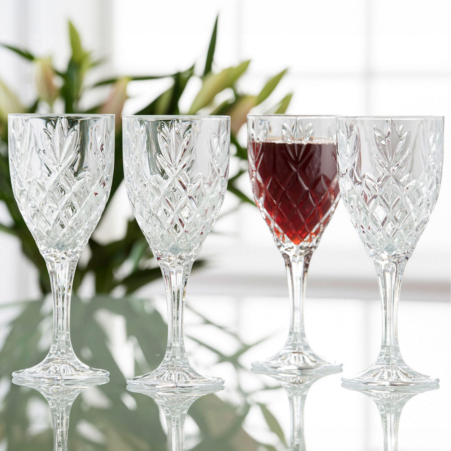 Four crystal wine glasses on a reflective surface, one filled with red wine, with a blurred floral background