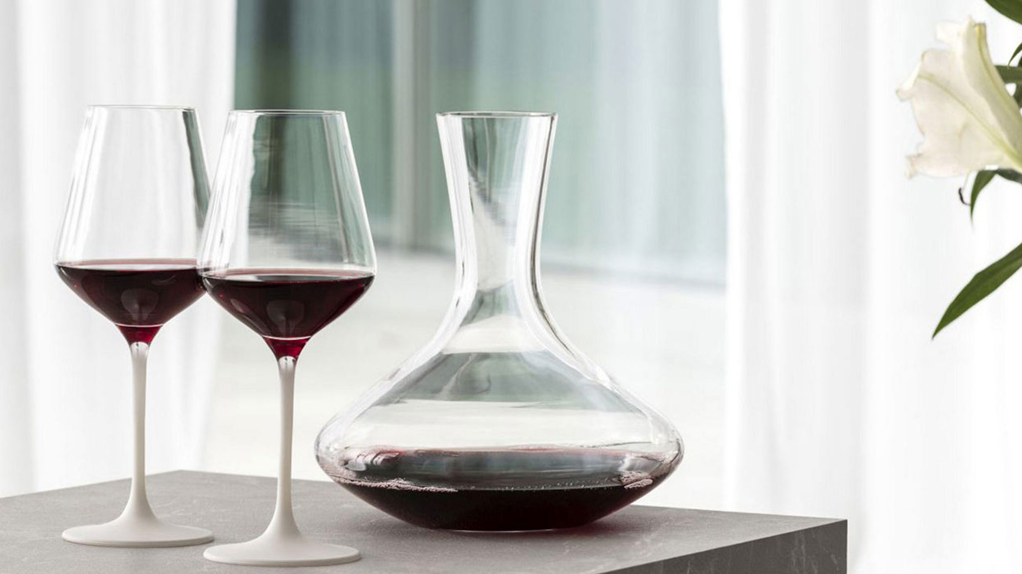 Two wine glasses, half filled with red wine and a decanter on a gray stone surface, with white curtains and lilies in the background.