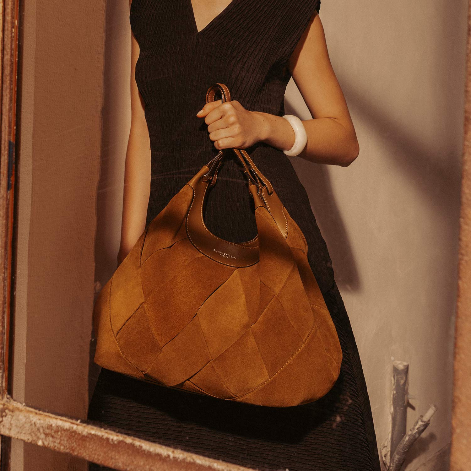 A woman in a dark dress holds a large, brown bag, standing by a rustic window frame.