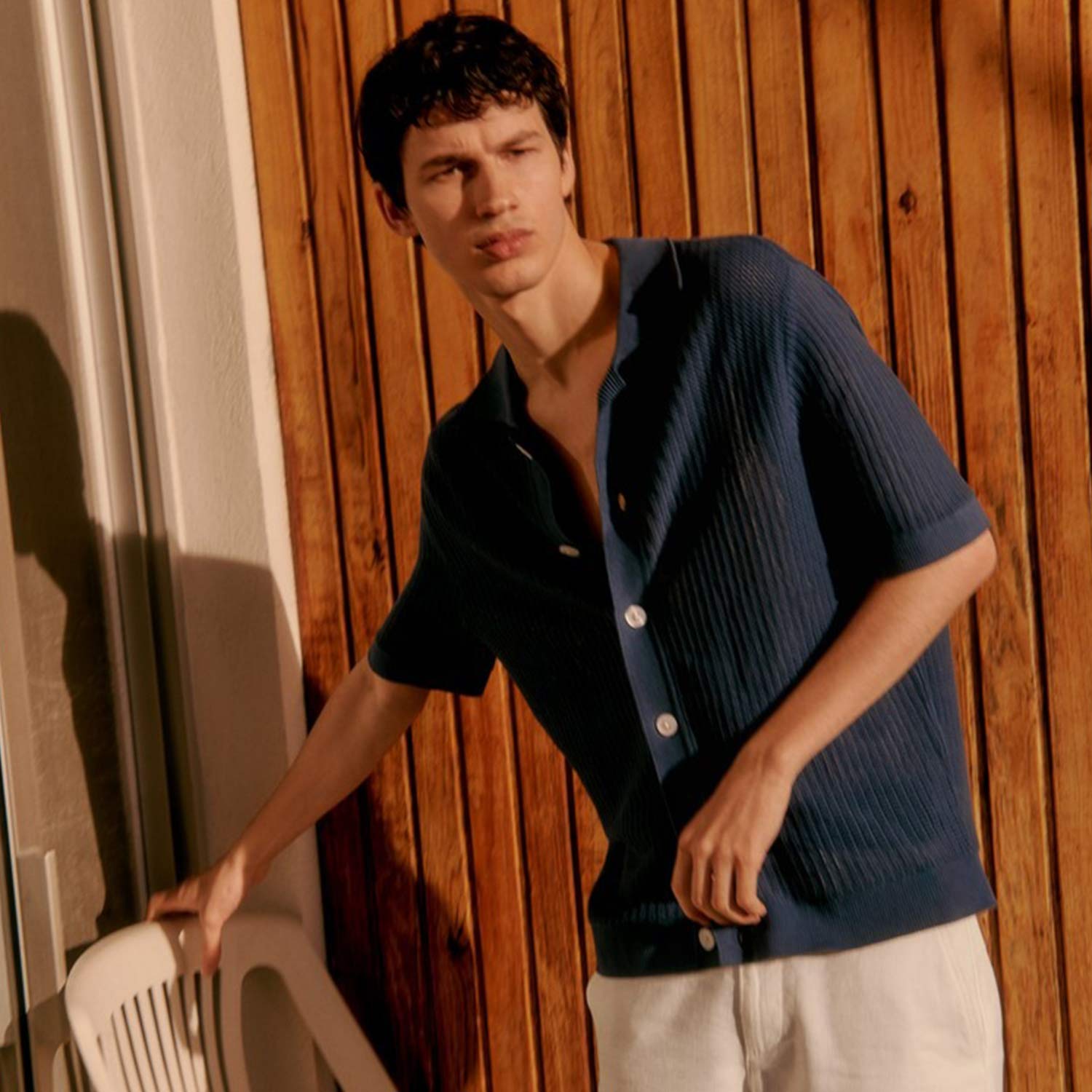 A person in a blue shirt and white shorts stands next to a white chair, set against a wooden backdrop.
