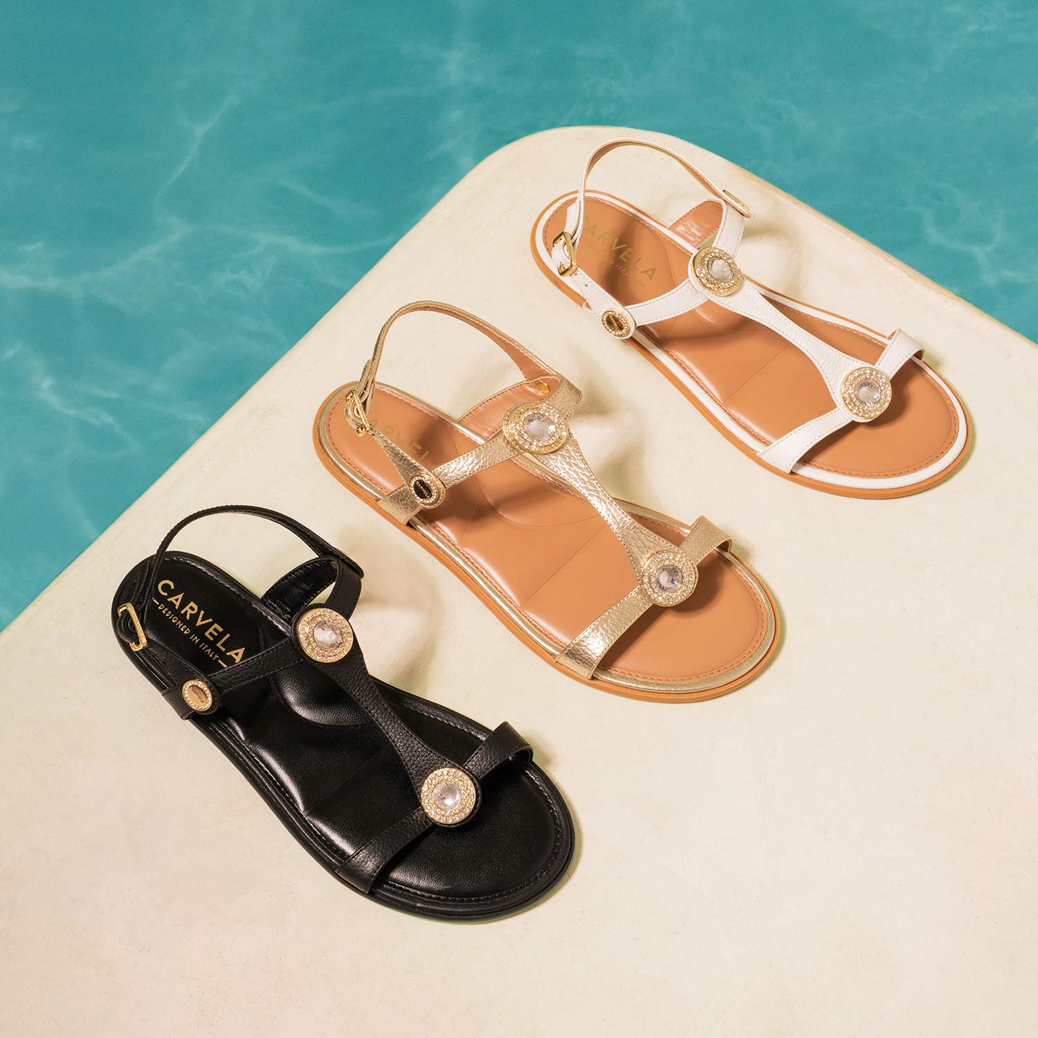 Three sandals in black, gold, and white are displayed on a cream surface beside a pool.