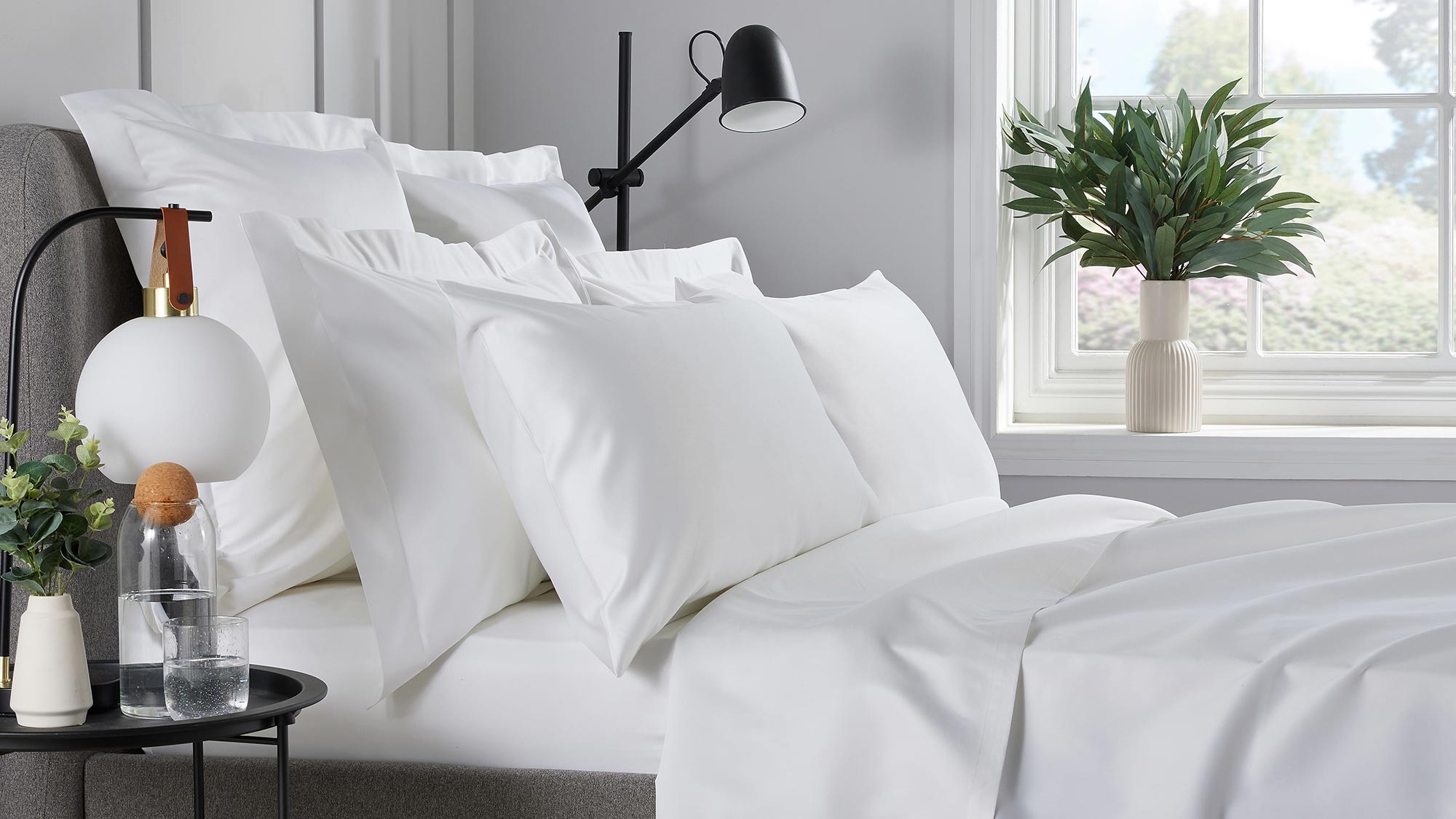 Modern bedroom with white bedding, black bedside lamp, potted plant, and minimalist decor on window sill