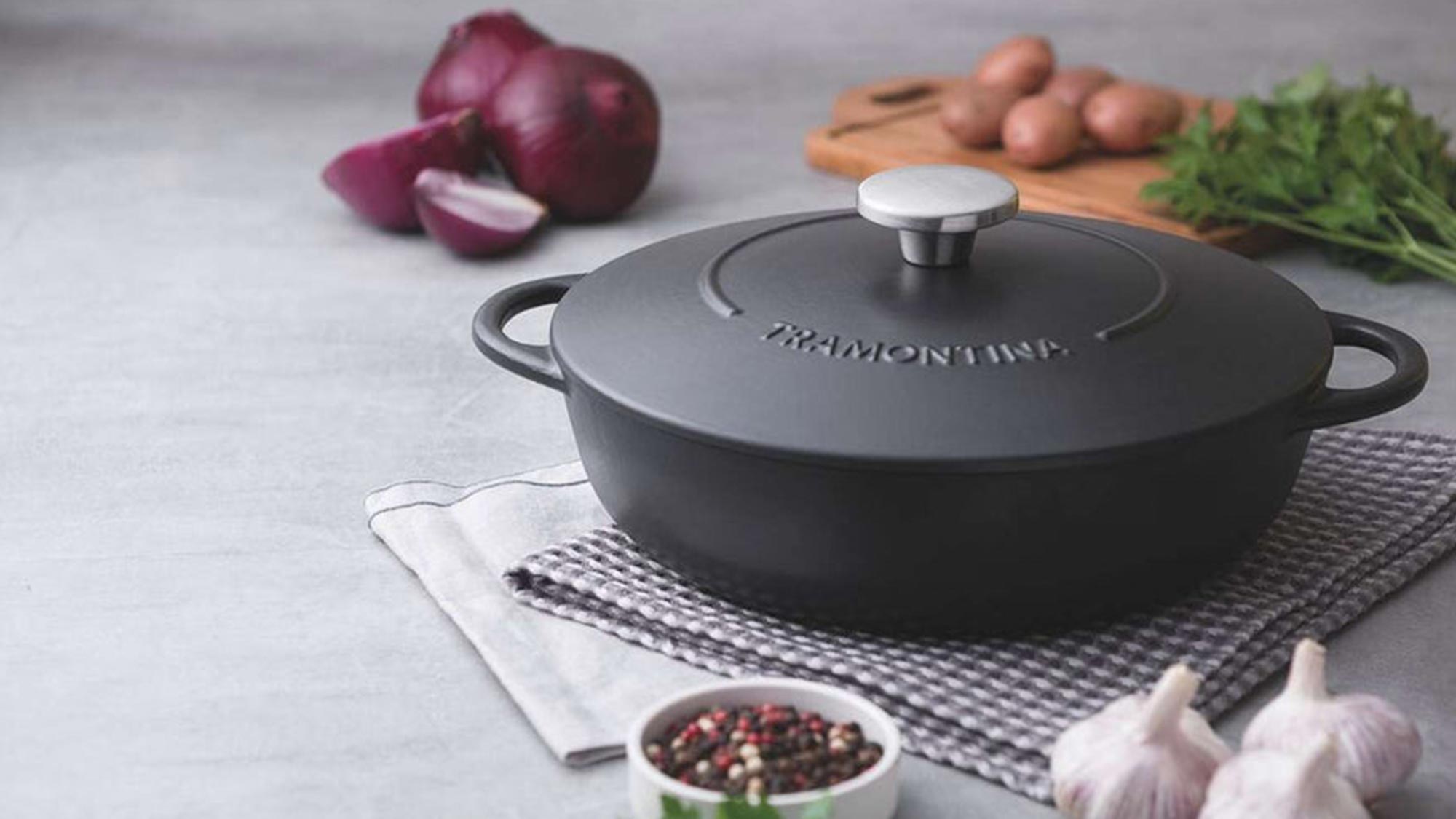 Black cast iron casserole dish on textured mat surrounded by fresh vegetables, herbs, and spices on countertop