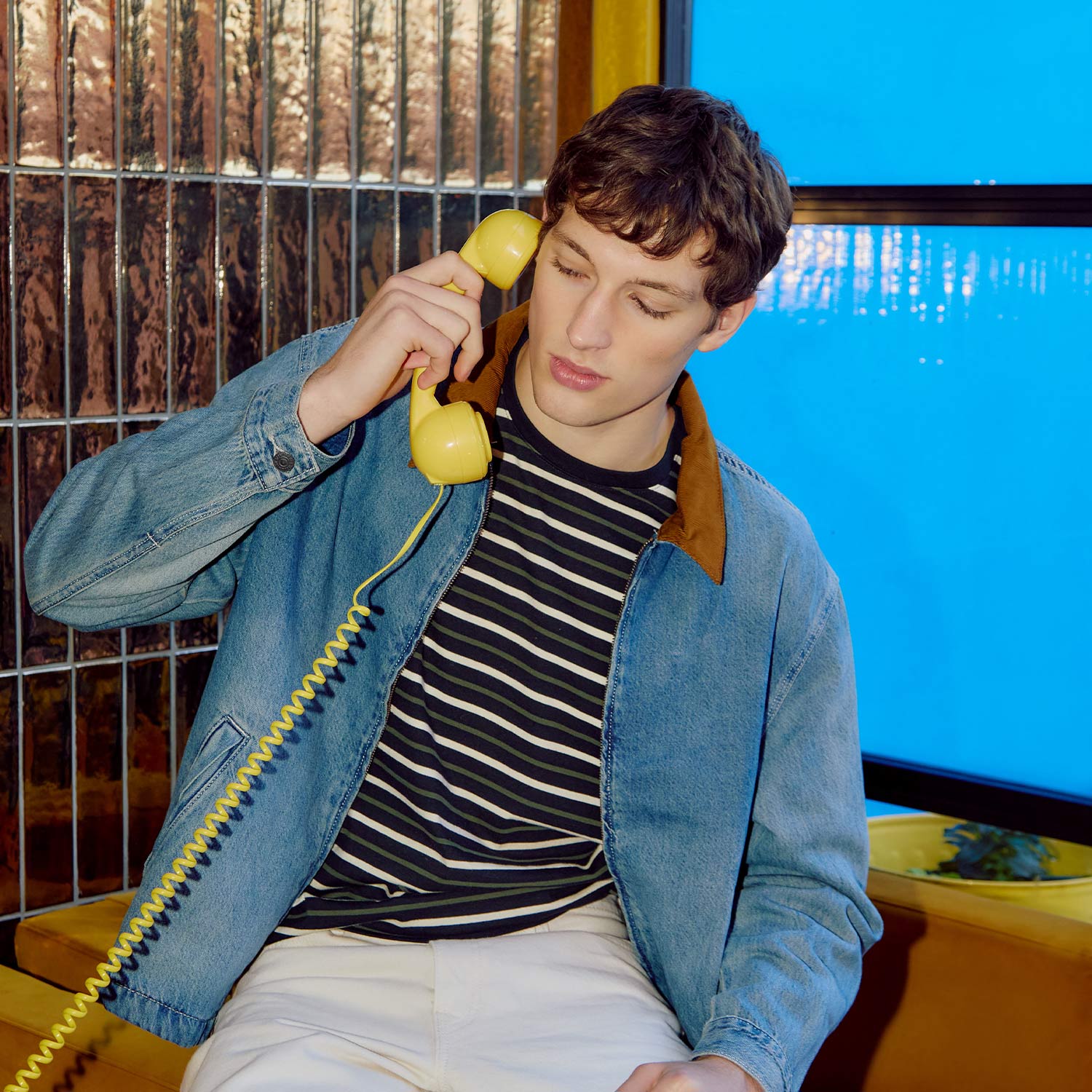 Young man wearing light blue denim jacket with brown collar, striped top, holding yellow rotary telephone handset against ear