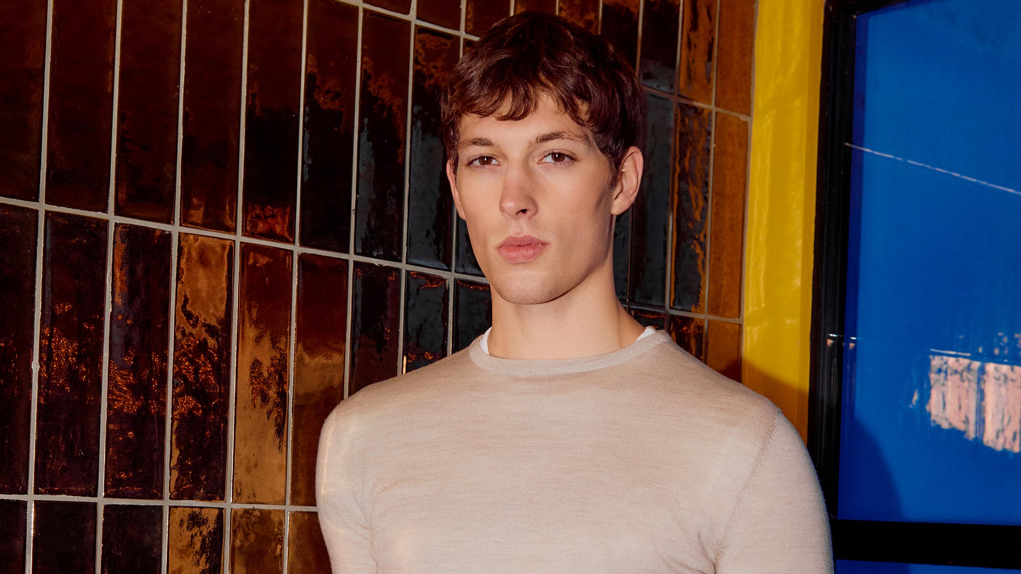 Man wearing beige crew neck knit jumper against textured rust and metallic tile backdrop