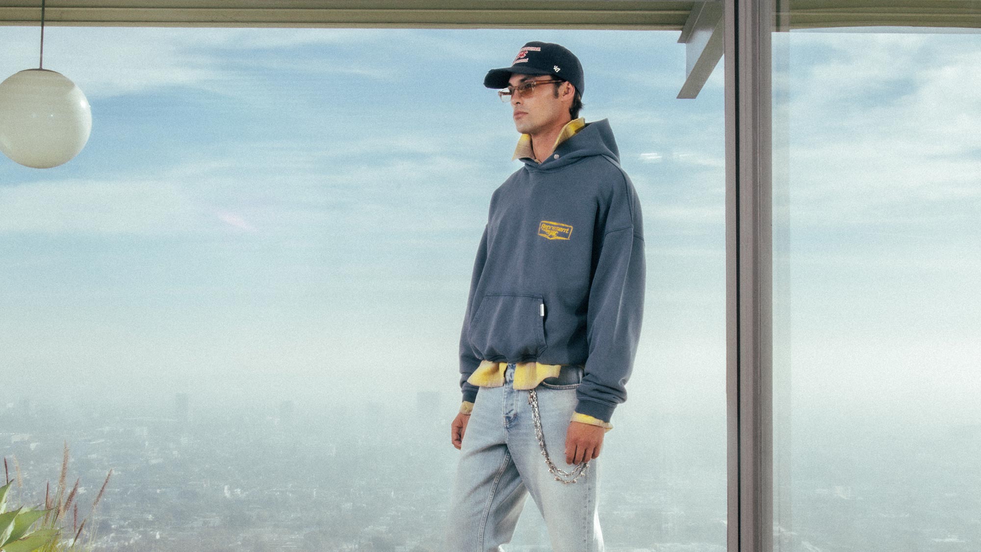 Man wearing black and yellow hoodie, light blue jeans with chain detail, and black baseball cap, standing by window with city skyline view