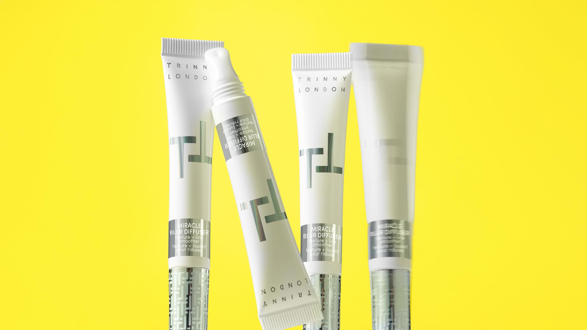 Trinny London Miracle Blur Diffuser products in white and grey tubes against bright yellow background
