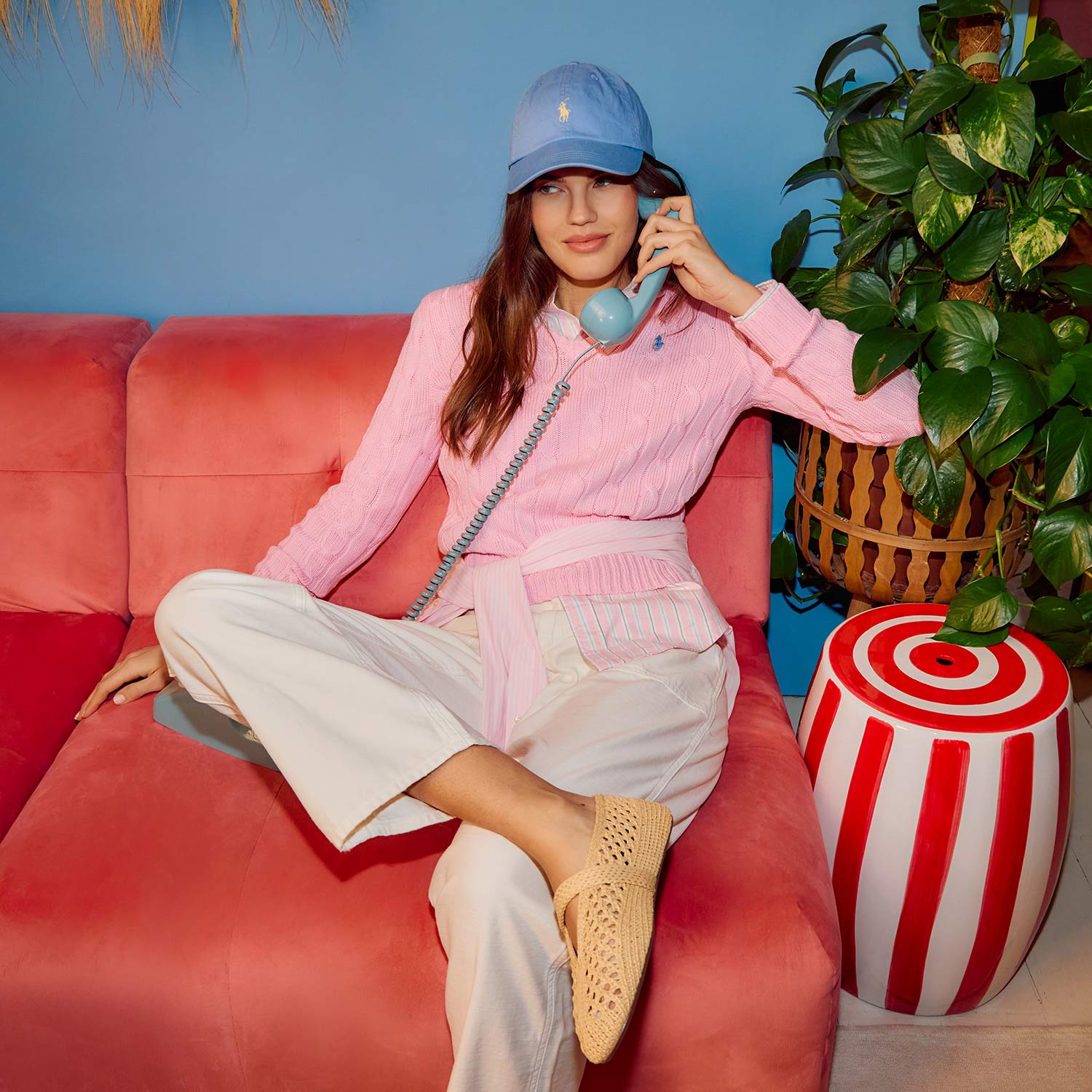 Woman in pink shirt and white trousers on red sofa wearing blue cap, holding telephone