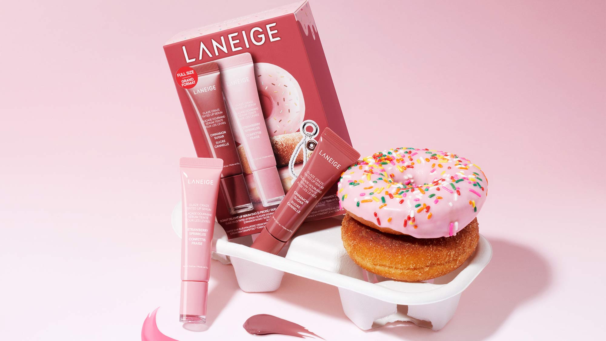 Laneige lip balm set with pink packaging next to a pink frosted doughnut with sprinkles. The set includes three tubes with different shades of pink balm. The items are displayed on a white stand against a pink backdrop, with smears of pink balm in front.