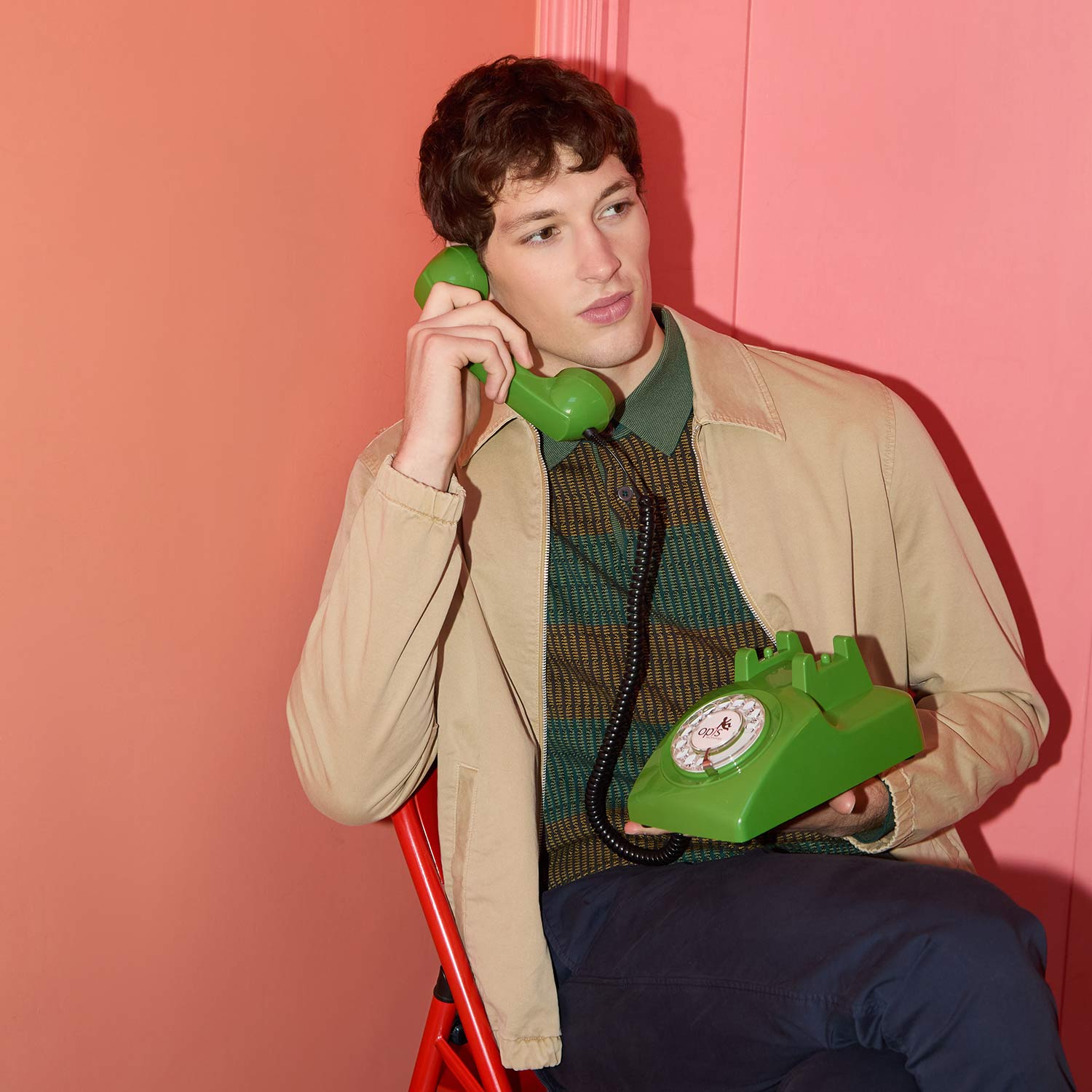 A person in a beige jacket is sitting and holding a green rotary phone against a pink-orange wall.