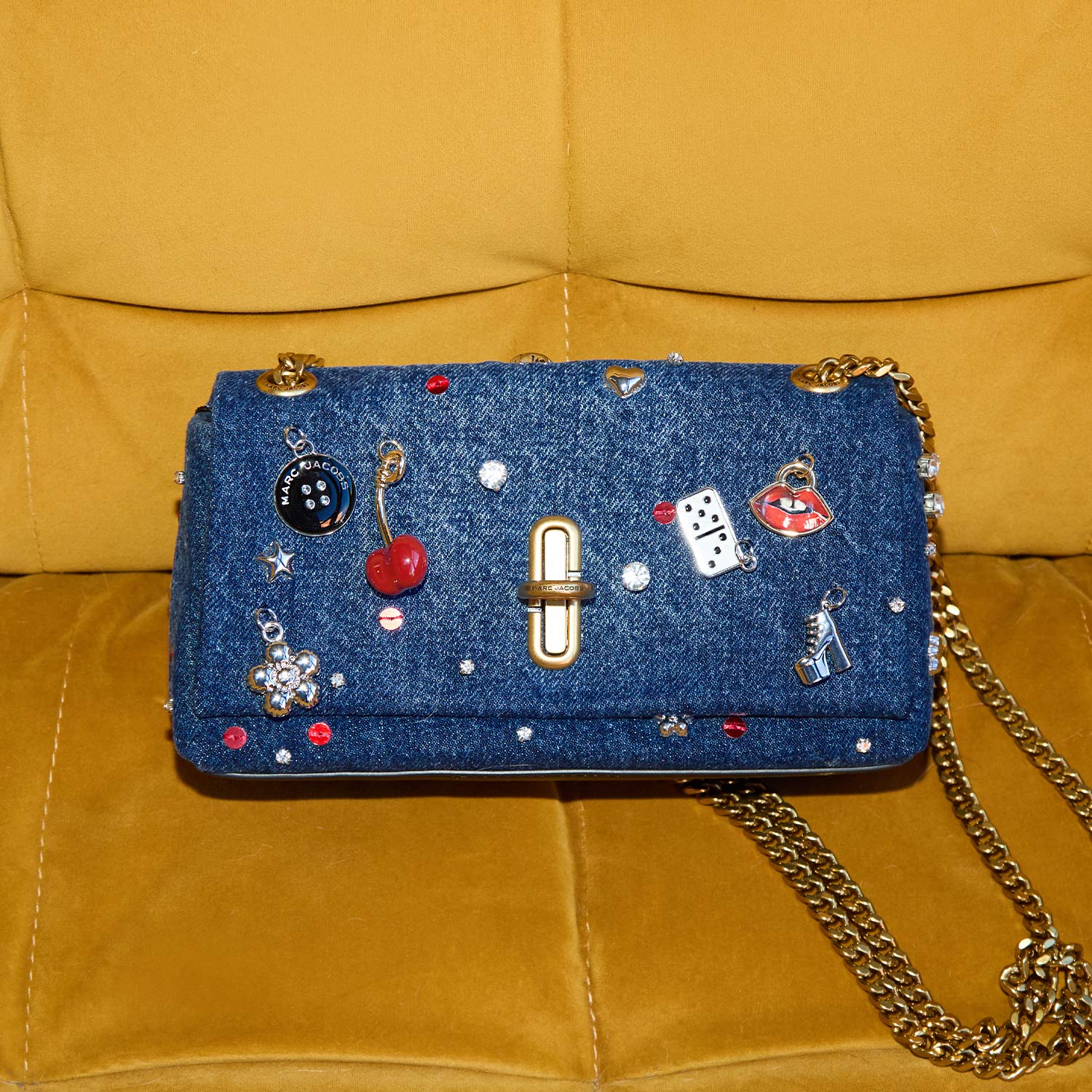 Blue denim crossbody bag with gold chain strap and decorative charms including cherries, dice and lips on yellow quilted sofa background