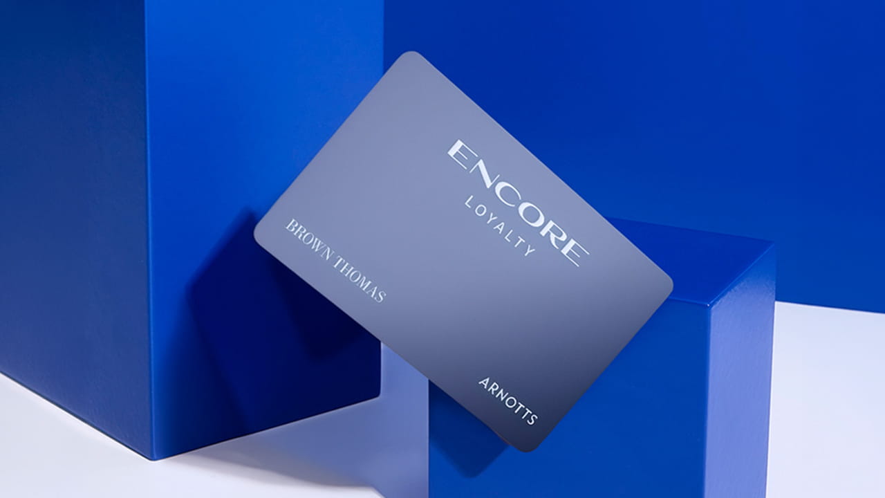A sleek loyalty card with 'ENCORE LOYALTY' text, partially shadowed, rests at an angle against a blue geometric backdrop with 'BROWN THOMAS' and 'ARNOTTS' printed on it.