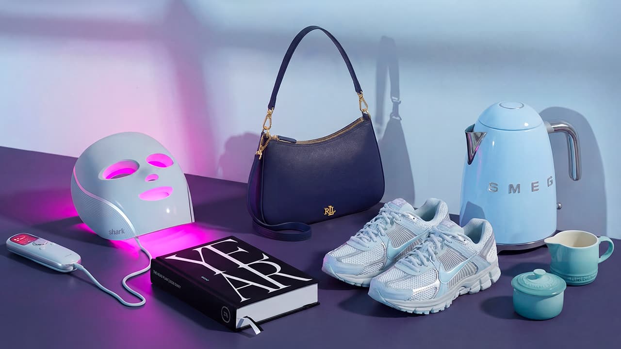 A collection of items including a light therapy mask, a book, a navy handbag, sneakers, a blue kettle, and teacups on a purple surface.