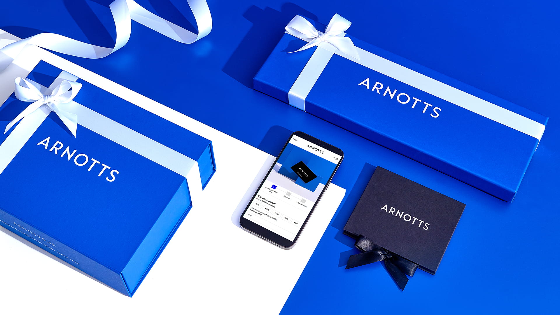 Blue gift boxes with white ribbons are labelled 'Arnotts' and arranged on a blue and white surface. A smartphone displays an online shopping page. A smaller black box with 'Arnotts' and a black ribbon accompanies the arrangement.