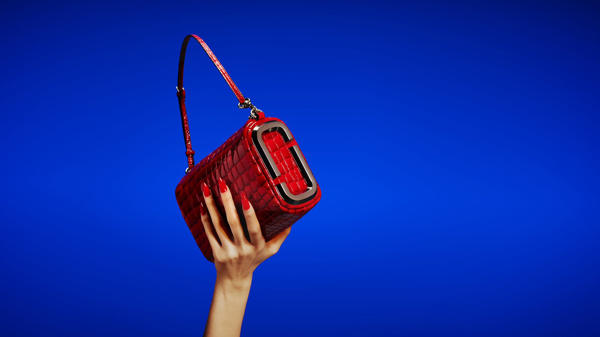Hand holding red quilted crossbody bag with gold hardware against bright blue background.