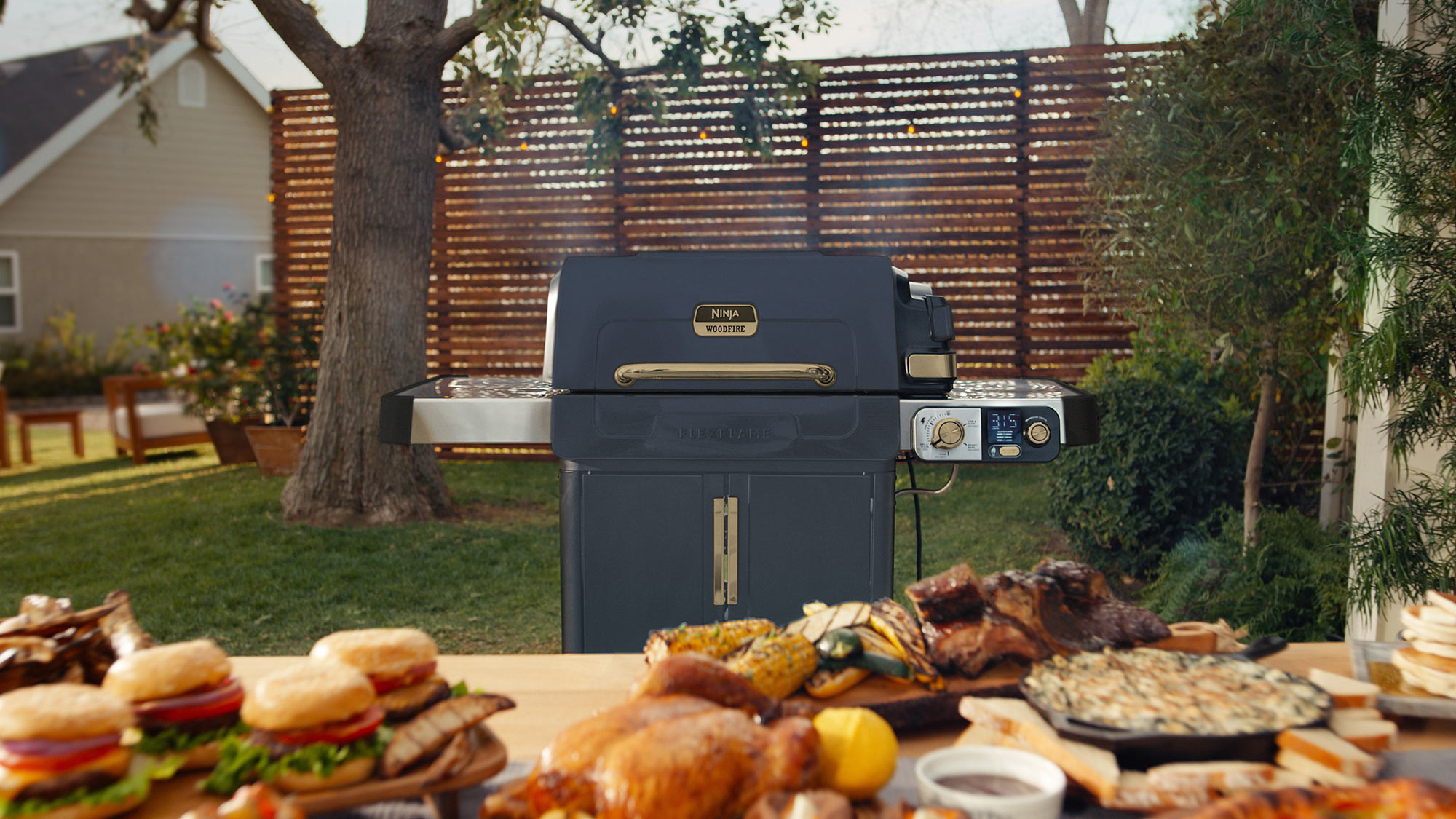 Ninja Woodfire barbecue on garden patio with cooked food and prepared dishes on table, ready for outdoor entertaining