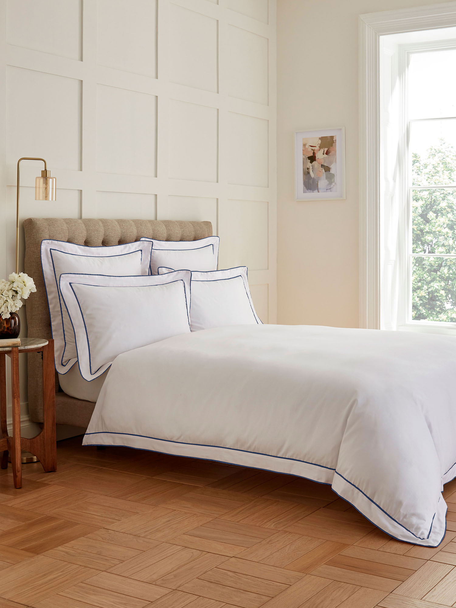 A bright bedroom with a neatly made bed, wooden floor, white paneled walls, a side table with flowers, and a large window.