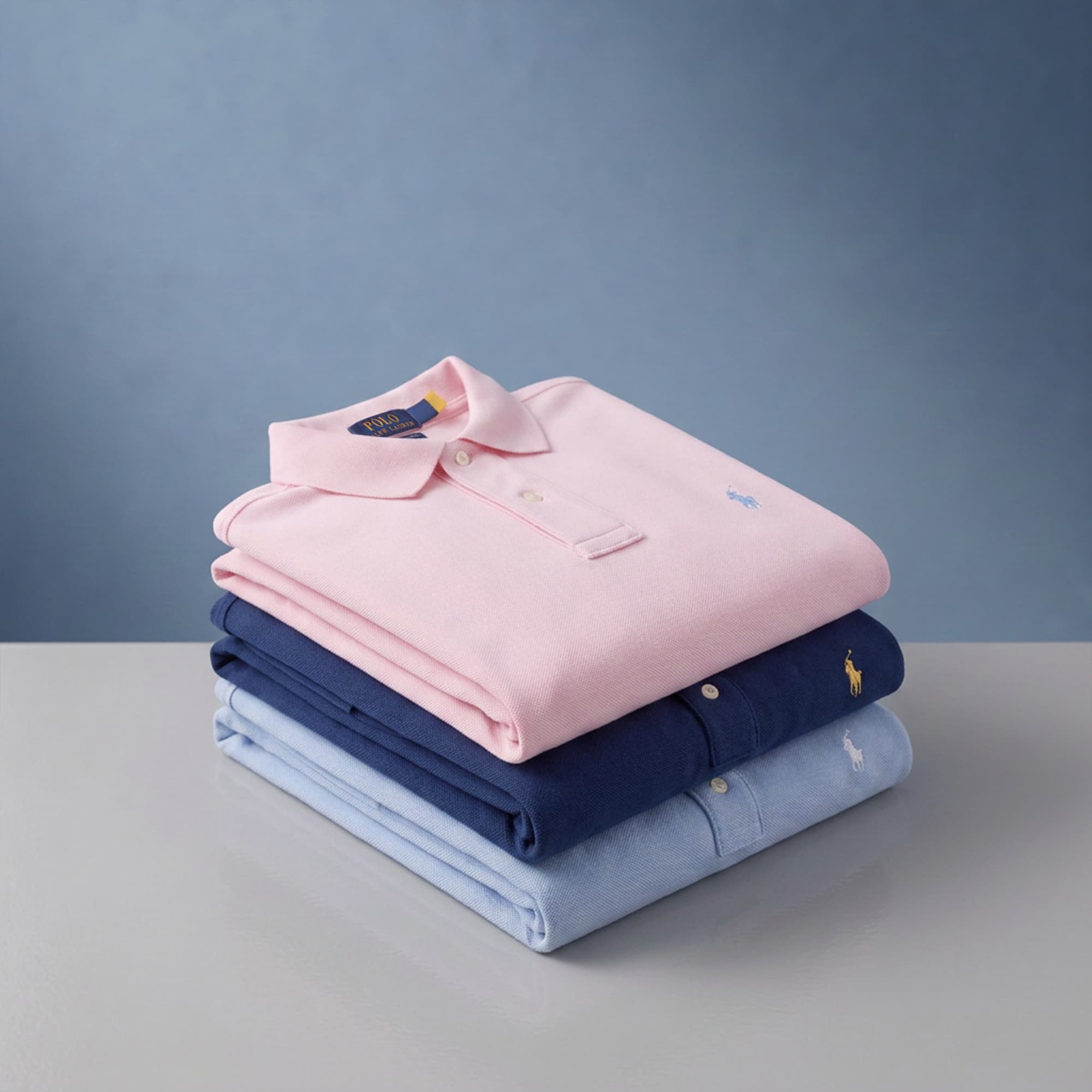 Three folded polo shirts are stacked on a grey surface against a plain background. The shirts are in pastel pink, navy blue, and light blue, each featuring a small embroidered logo on the chest.