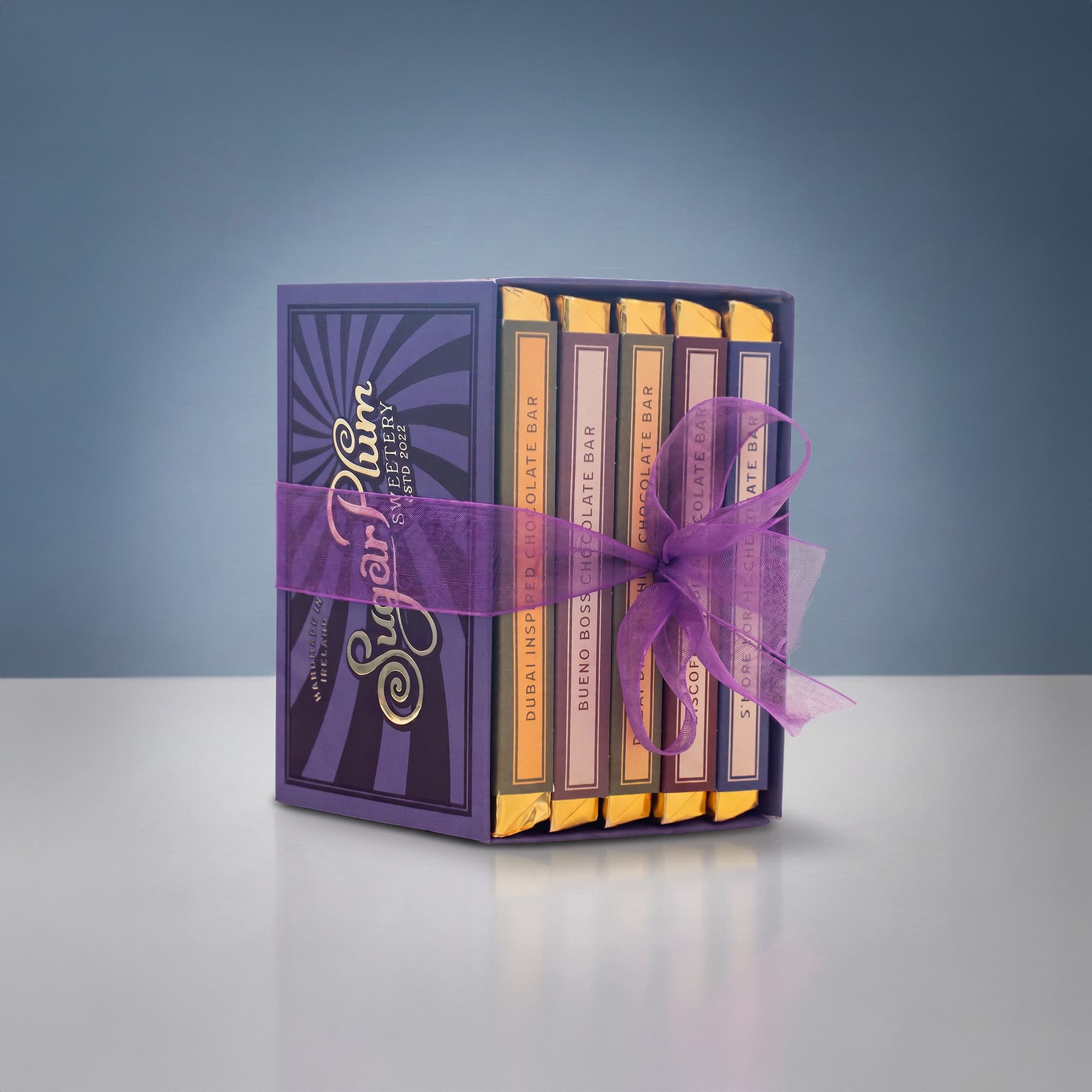 A set of five luxury chocolate bars in purple packaging, neatly tied with a sheer purple ribbon. The bars are arranged vertically and feature elegant gold and pink labels, set against a gradient blue background.