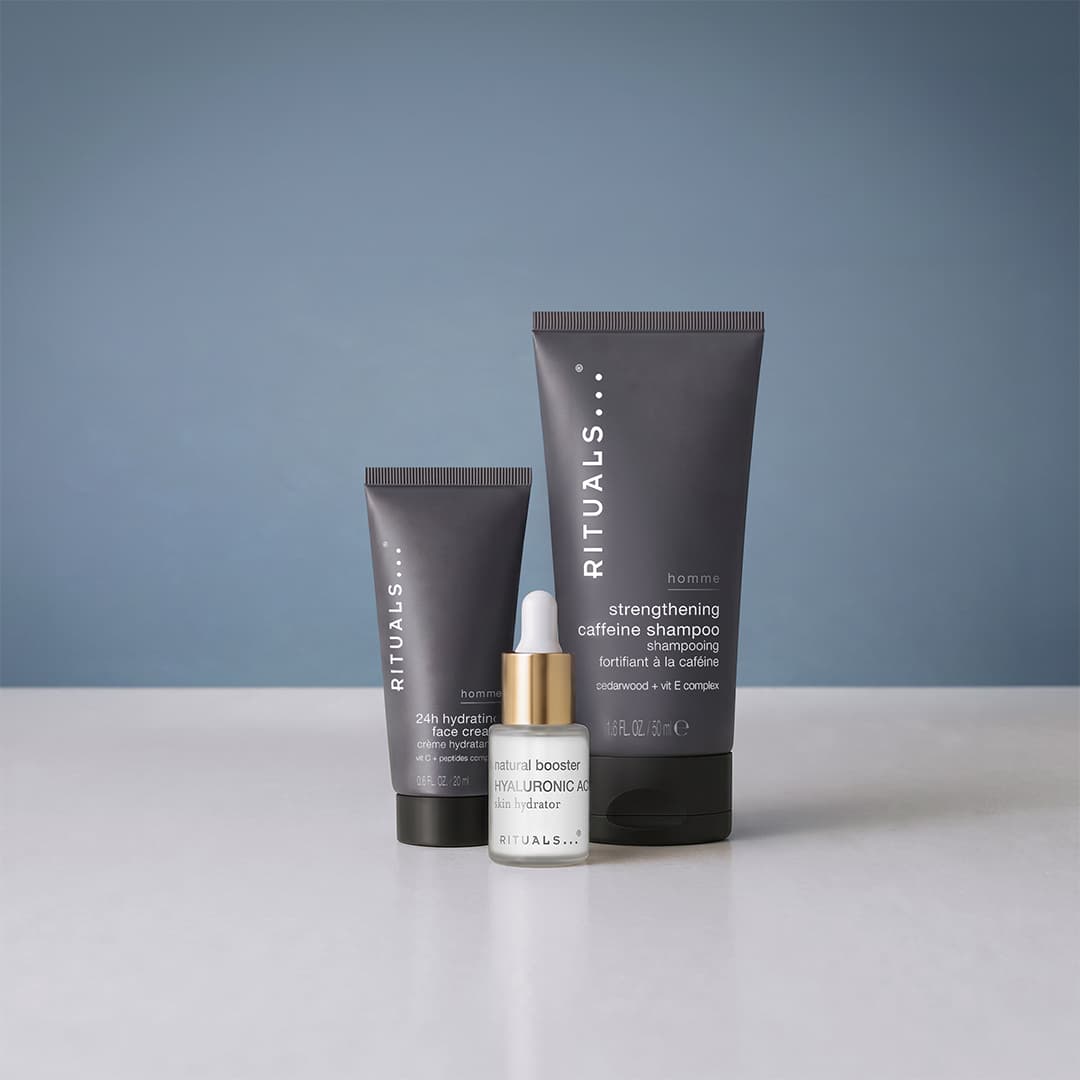 Three Rituals skincare products against a plain blue background. A tube of strengthening caffeine shampoo is on the right, a smaller tube of 24h hydrating face cream is on the left, and a bottle of natural booster serum is in the foreground.